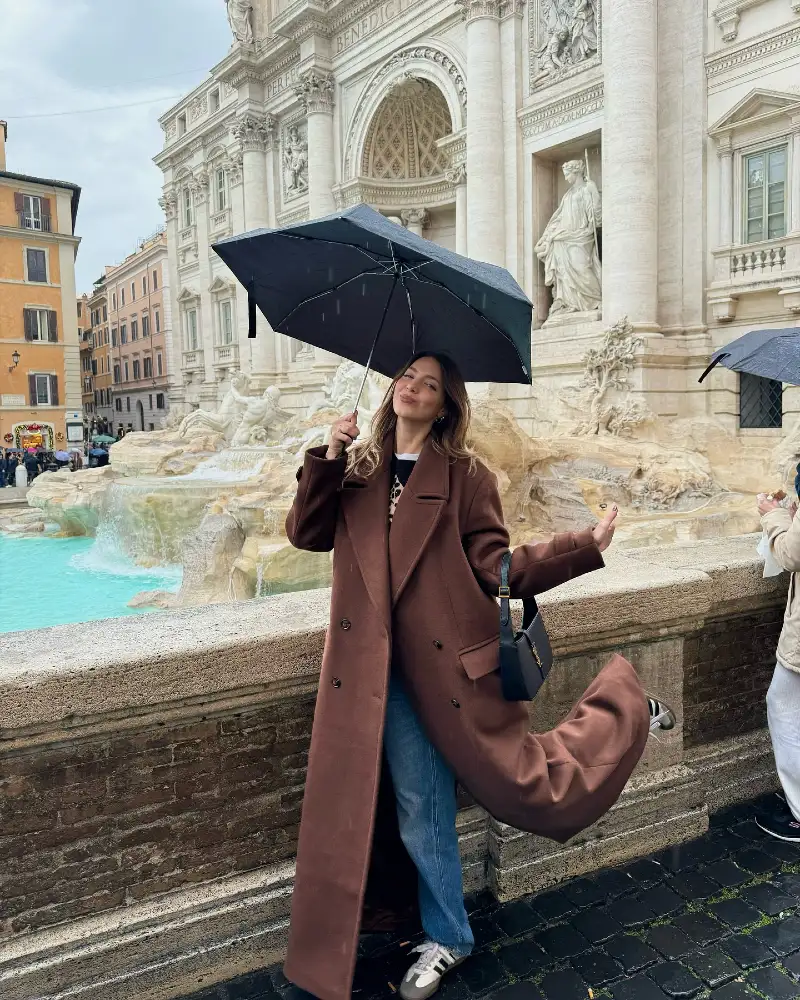 Long Brown Wool Coat with Classic Denim, Sneakers, and Black Umbrella in Front of Historic Fountain